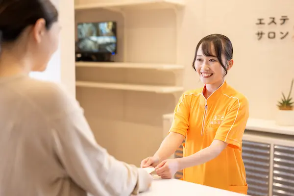 エステサロンの受付で、笑顔のスタッフがお客様に書類やカードを手渡している様子